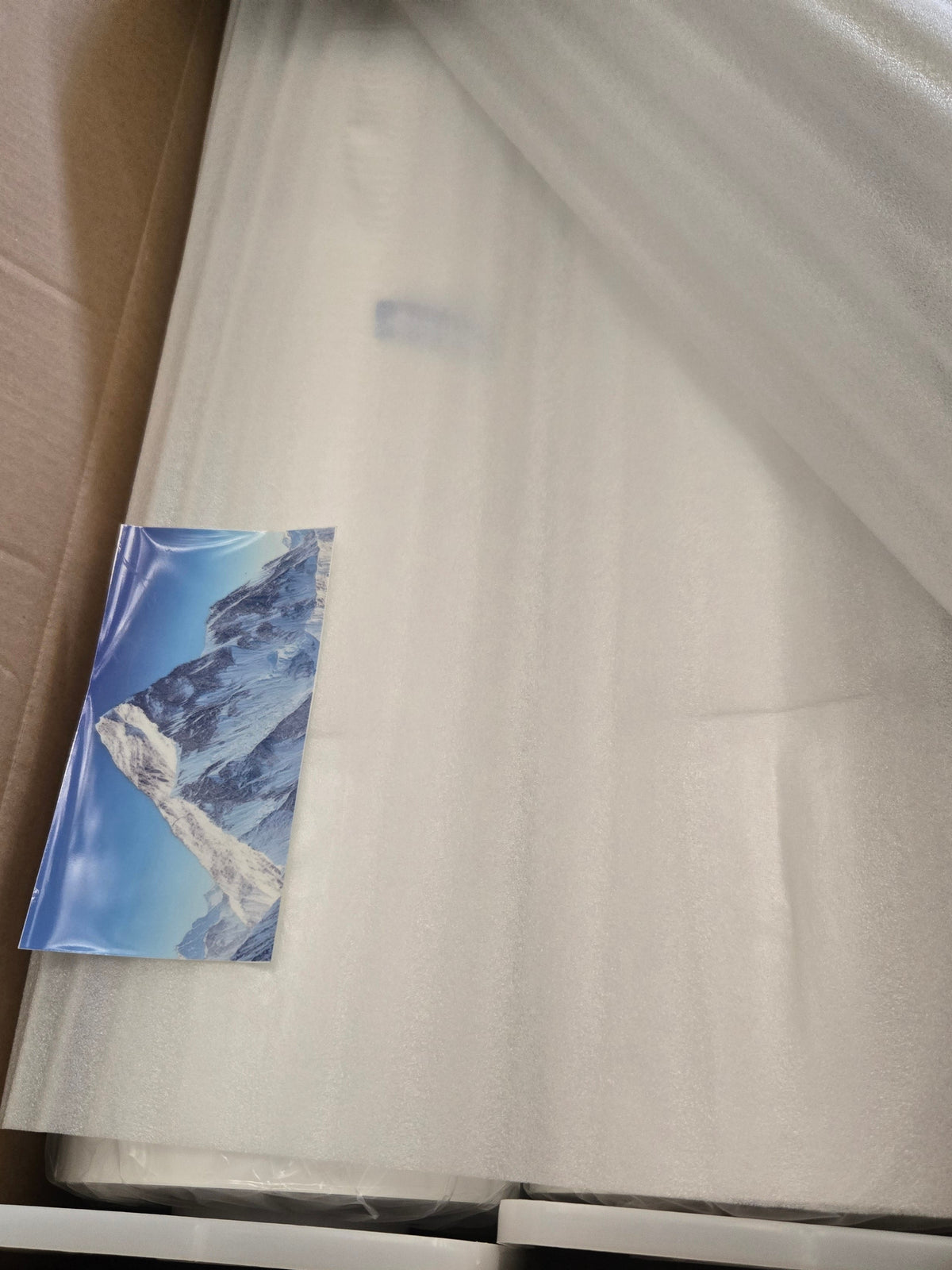 A large white rectangular item wrapped in protective foam and plastic inside a brown cardboard box, with a small printed card featuring a blue mountain scene tucked near the left side. A hand is visible at the bottom right.
